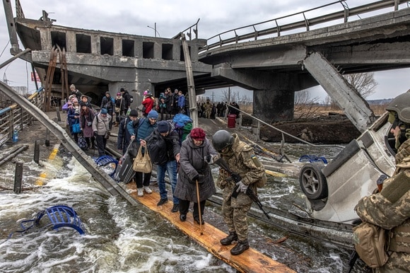 Soldier helping civilians across a plank crossing a river.